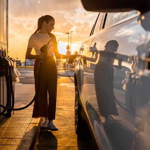 Beautiful young woman refueling gas tank while standing next to car at fuel pump during sunset