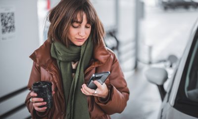 Portrait of a young woman with phone and coffee to go standing at gas station outdoors, waiting for the car to refuel