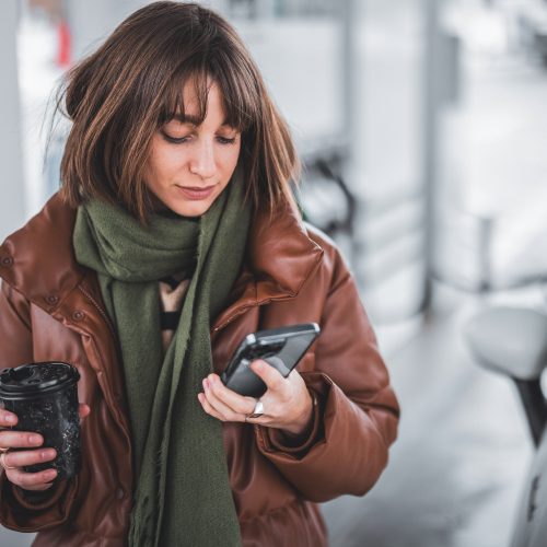 Portrait of a young woman with phone and coffee to go standing at gas station outdoors, waiting for the car to refuel