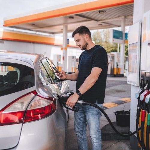 Bearded man refuelling car on gas station and looking into his smartphone. Man compares fuel prices . High quality photo